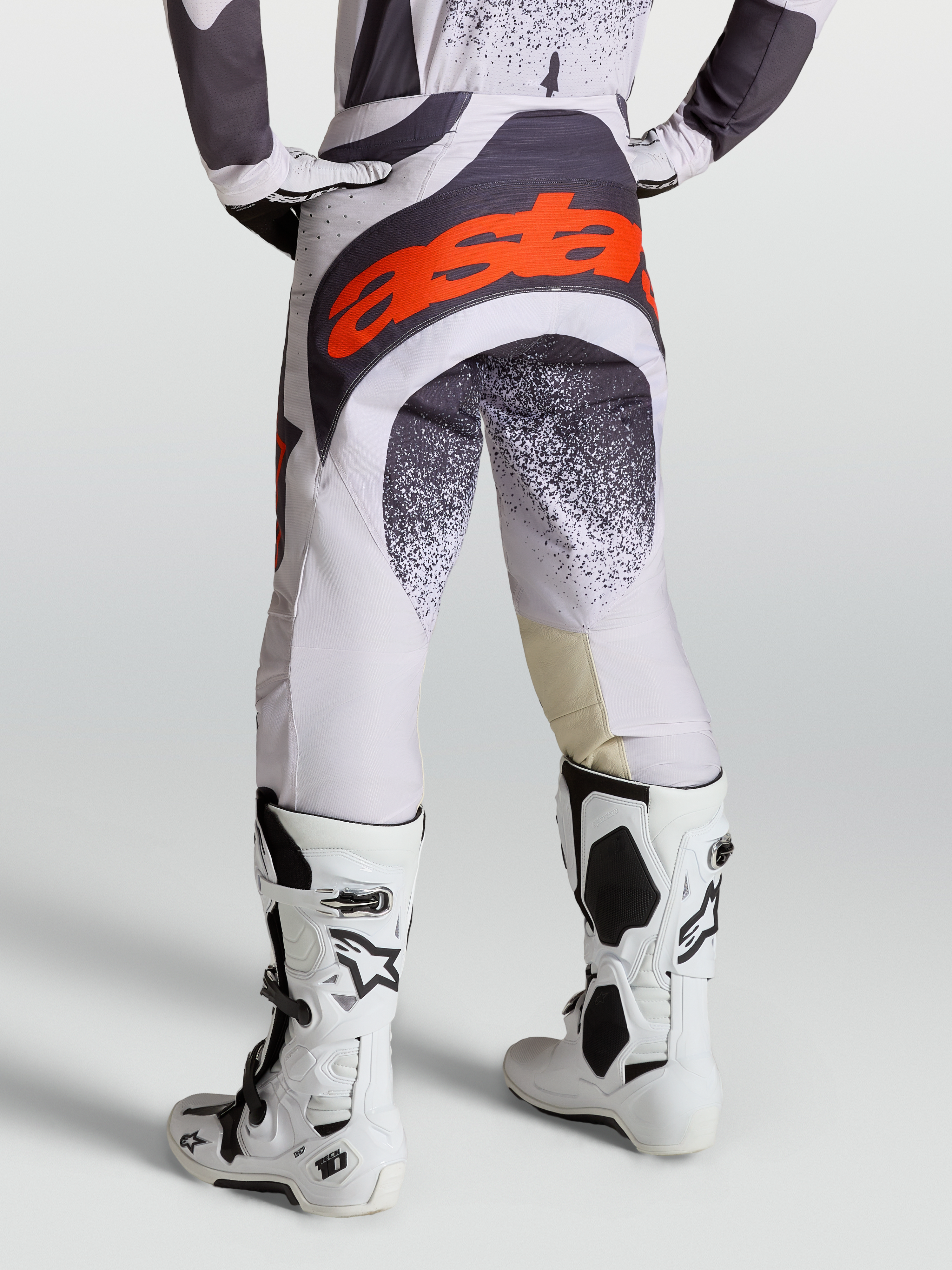Alpinestars Techstar Swan broek voor heren, motocrossracebroek in licht lila en donkergrijs met rood fluo accenten, gedragen met witte Tech 10 motorlaarzen, professionele off-road motoruitrusting.