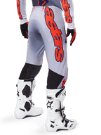Alpinestars 2025 Supertech Lipan crossbroek, motorcrossbroek, grijs en fluoranje, gedragen met bijpassende jersey en witte laarzen, achteraanzicht met grote oranje Alpinestars grafische branding, zwarte stretchpanelen, professionele motorcrossuitrusting