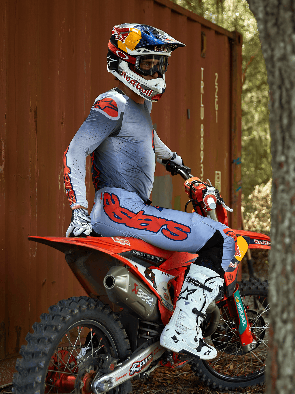 Motorcrossrijder zittend op een rode crossmotor, gekleed in de Alpinestars 2025 Supertech Lipan Gear Set in Grijs en Oranje Fluo, lichtblauwe broek met opvallend oranje Astars-logo, bijpassende jersey, witte motorcrosslaarzen en een Red Bull-helm, gecoördineerde professionele race-uitrusting