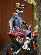 Motorcrossrijder zittend op een rode crossmotor, gekleed in de Alpinestars 2025 Supertech Lipan Gear Set in Grijs en Oranje Fluo, lichtblauwe broek met opvallend oranje Astars-logo, bijpassende jersey, witte motorcrosslaarzen en een Red Bull-helm, gecoördineerde professionele race-uitrusting