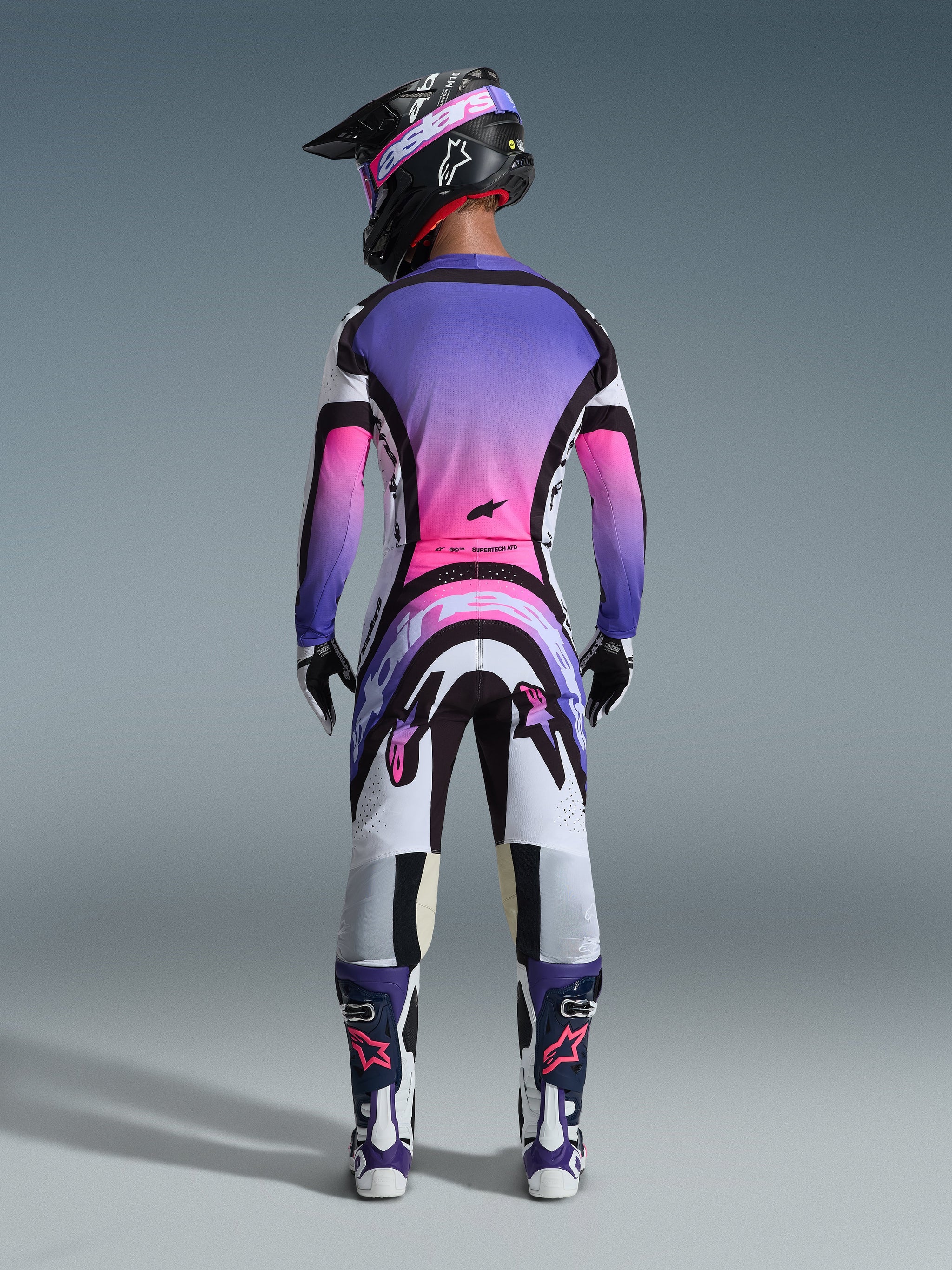Motocrossrijder van achteren gezien in volledige race-uitrusting, shirt met paars en roze kleurverloop, Alpinestars 2026 Supertech AFD Vista broek in White Purple Black, zwarte helm met paarse crossbril, witte en paarse laarzen, gecoördineerde off-road racekit.
