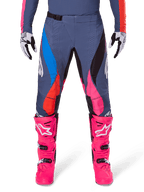 Alpinestars 2025 Techstar Dreem broek, motorcross broek, grijs multicolor met blauwe, roze en oranje accenten, gedragen met felroze laarzen en wit-zwarte handschoenen, voorzien van lasergesneden perforaties op de dijen en een magnetische taillesluiting, atletische race-fit uitrusting voor off-road competitie