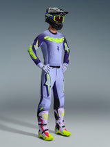Alpinestars 2026 Supertech Scenz broek, motorcrossbroek, paars en geel fluo, gedragen door een rijder in een volledige race-outfit met bijpassende jersey met kleurverloop, lila handschoenen en bijbehorende geel-paarse laarzen, voorzien van een zwarte helm en bril.