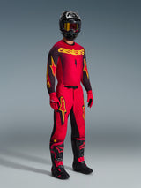 Alpinestars 2026 Supertech Scenz broek, motorcrossbroek, rood, zwart en geel, gedragen door een rijder in een volledige race-uitrusting met bijbehorende jersey en laarzen, opvallende geel-zwarte sterlogo's en een atletische pasvorm.