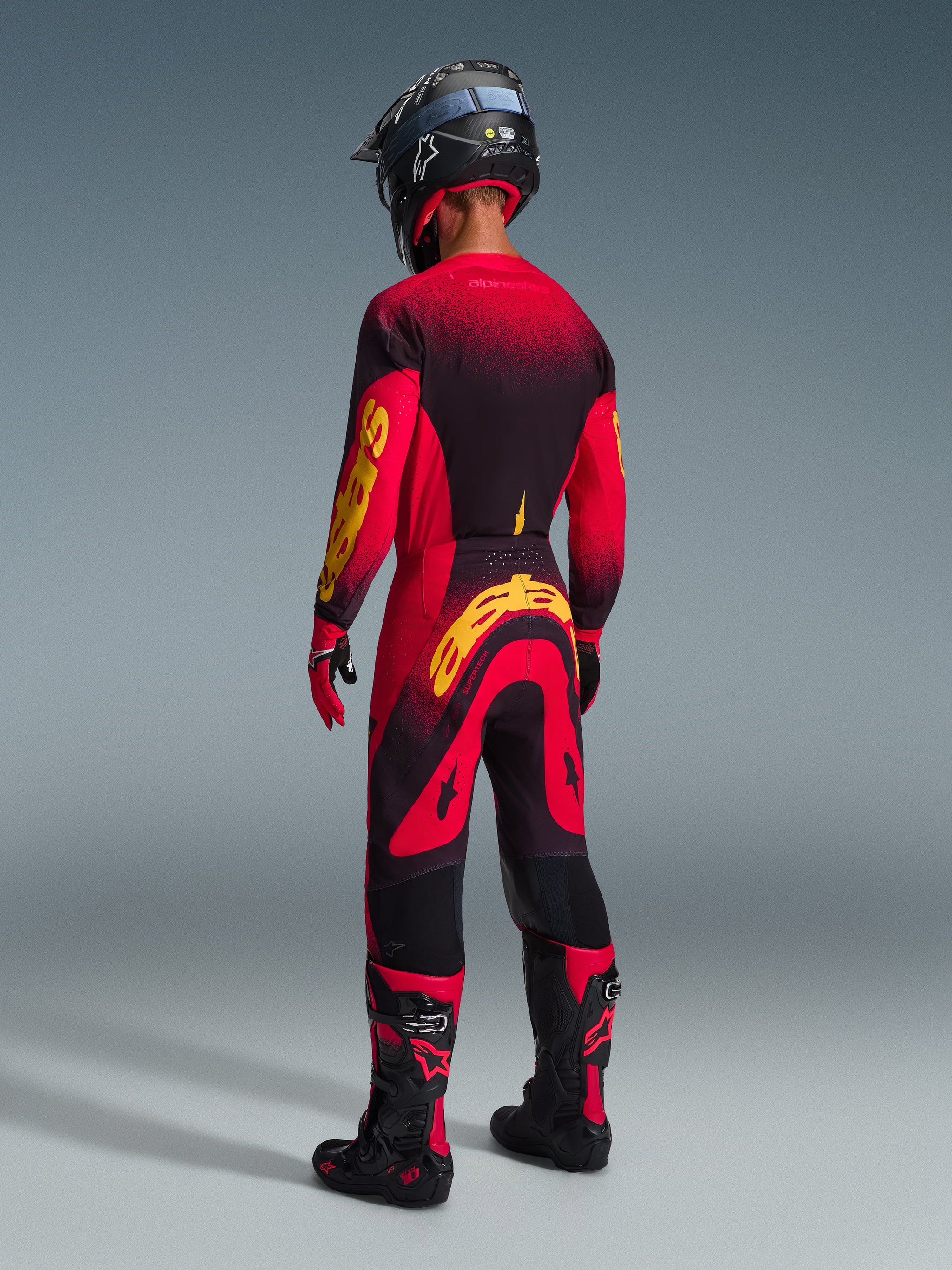 Motorcrosser van achteren gezien in Alpinestars 2026 Supertech Scenz broek, rood zwart geel, bijpassende jersey met opvallende gele branding, zwarte en rode motorcrosslaarzen, zwarte helm, complete professionele race-uitrusting voor off-road prestaties.