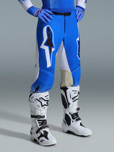 Alpinestars 2026 Supertech Scenz broek, motorcrossbroek, Ucla blauw en wit, gedragen met witte en zwarte laarzen en blauwe handschoenen, voorzien van grote grafische logo's op de dijbenen en witte panelen, atletische pasvorm ontworpen voor competitief motorcrossen