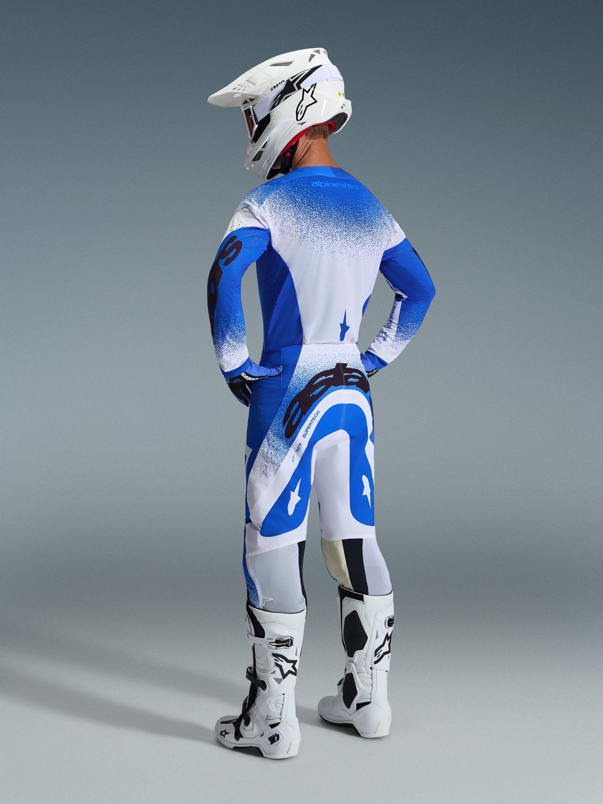 Motorcrosser vanaf de achterzijde in de 2026 Supertech Scenz broek en jersey in Ucla Blue White, witte helm met Alpinestars-logo, witte en zwarte motorcrosslaarzen, professionele off-road race-uitrusting.