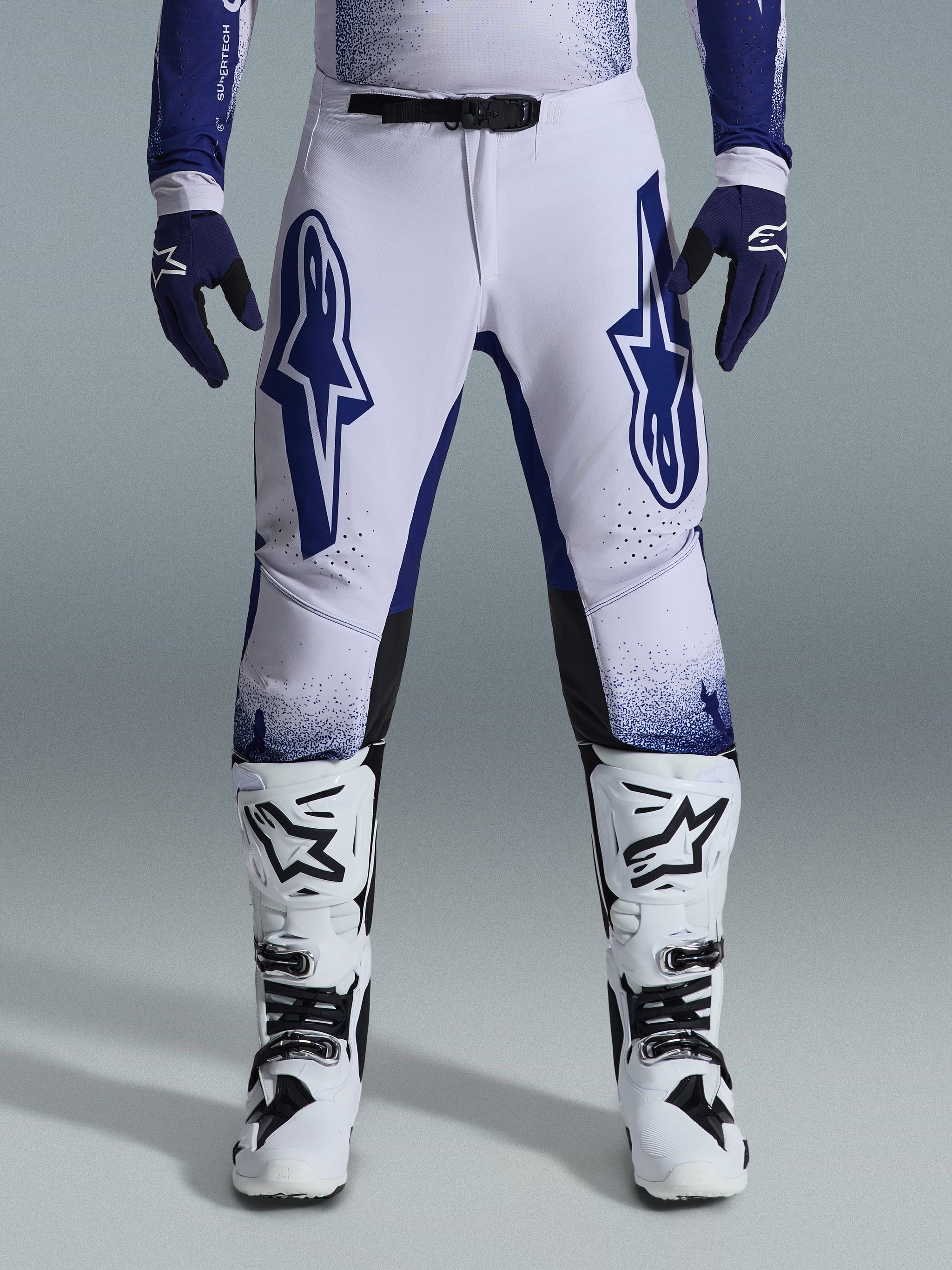 Alpinestars 2026 Supertech Scenz broek, motorcross broek, lichtgrijs en donkerblauw, gedragen met wit-zwarte motorcrosslaarzen en donkerblauwe handschoenen, voorzien van grote blauwe Alpinestars-logo's op de dijen, SX1 magnetische gesp en atletische pasvorm voor off-road racing