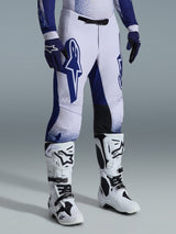 Alpinestars 2026 Supertech Scenz broek, motorcrossbroek, lichtgrijs en donkerblauw, gedragen met witte motorcrosslaarzen en blauwe handschoenen, voorzien van grote blauwe Alpinestars-logo's op de dijen, zwarte kniepanelen en een gestippeld grafisch ontwerp, professionele off-road race-uitrusting