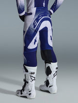 Alpinestars 2026 Supertech Scenz broek, motorcrossbroek, lichtgrijs en donkerblauw, achteraanzicht gedragen met bijpassende jersey en witte laarzen, opvallende blauwe graphics met gespikkelde accenten, sportieve atletische pasvorm voor off-road racen