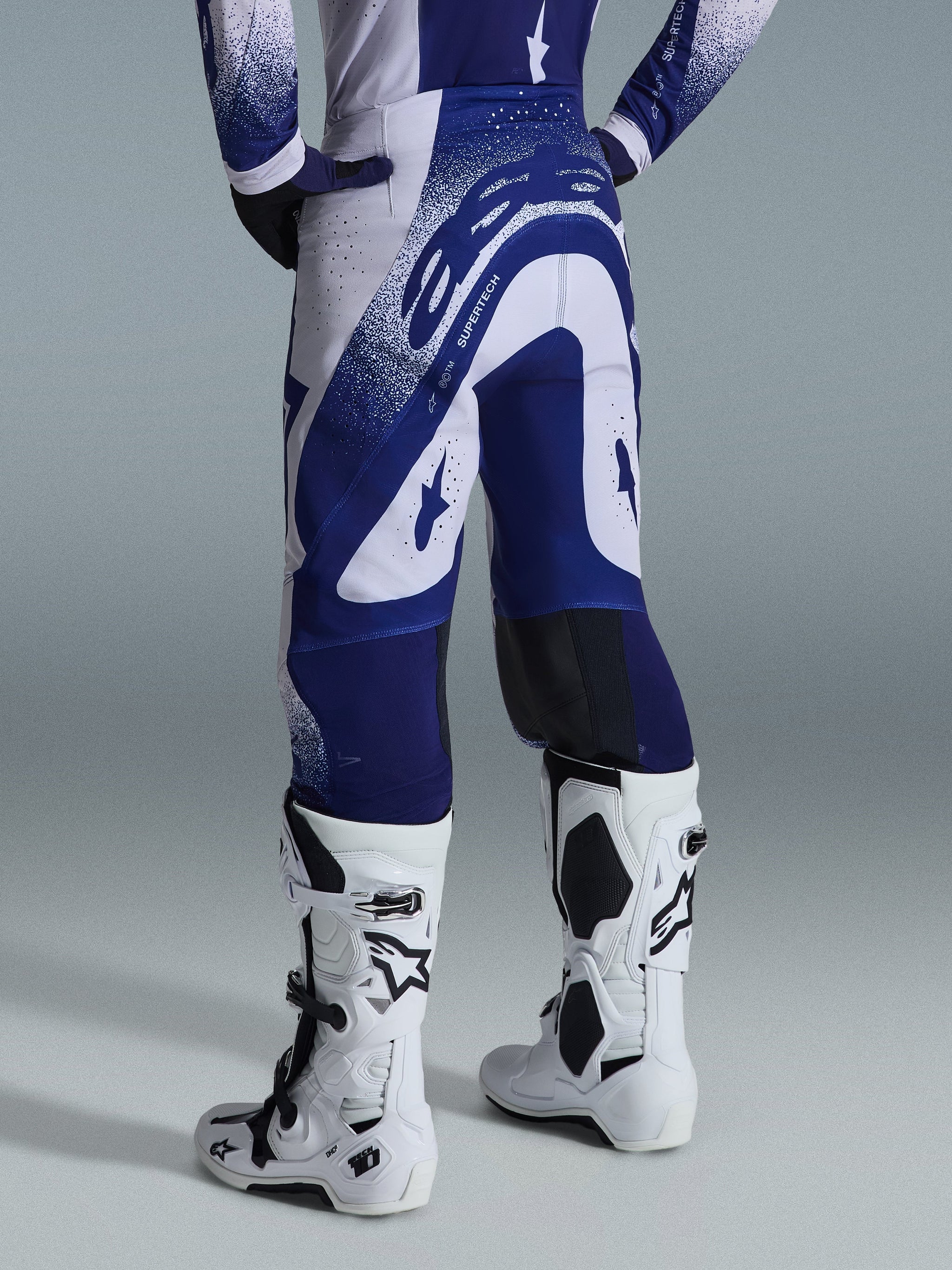 Alpinestars 2026 Supertech Scenz broek, motorcrossbroek, lichtgrijs en donkerblauw, achteraanzicht gedragen met bijpassende jersey en witte laarzen, opvallende blauwe graphics met gespikkelde accenten, sportieve atletische pasvorm voor off-road racen