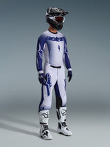 Alpinestars motorcrossrijder in 2026 Supertech Scenz broek, lichtgrijs en donkerblauw, bijpassende blauw-witte jersey met 'astars' branding en spray-paint effect, zwarte helm met witte goggle en witte en zwarte motorcrosslaarzen, gecoördineerde professionele off-road race-outfit.