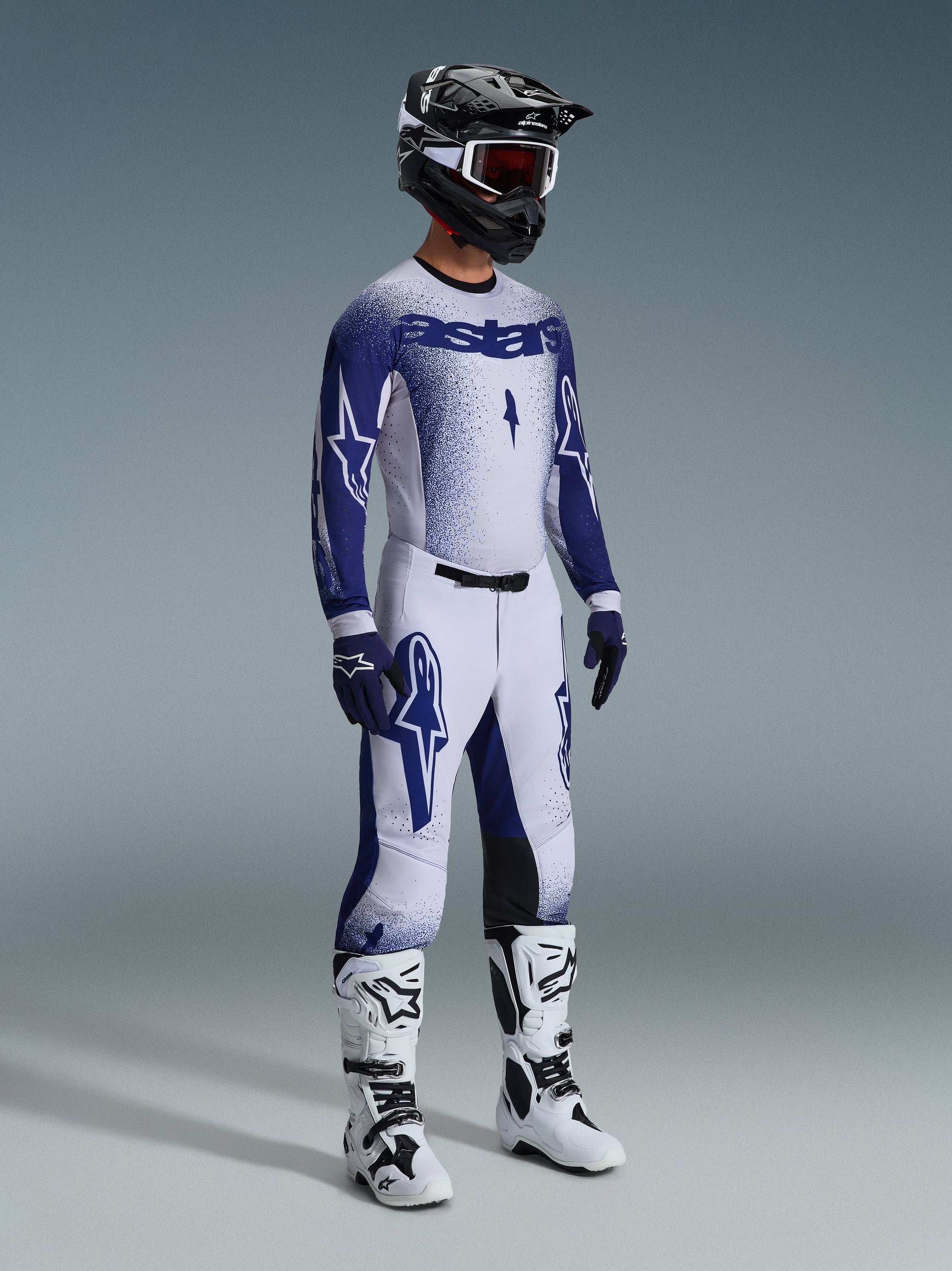 Alpinestars motorcrossrijder in 2026 Supertech Scenz broek, lichtgrijs en donkerblauw, bijpassende blauw-witte jersey met 'astars' branding en spray-paint effect, zwarte helm met witte goggle en witte en zwarte motorcrosslaarzen, gecoördineerde professionele off-road race-outfit.