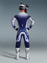 Alpinestars 2026 Supertech Scenz broek, motorcrossbroek, lichtgrijs en donkerblauw, achteraanzicht van een rijder in een bijpassende kledingset met gespikkelde kleurverloop-graphics, witte laarzen en zwarte helm, professionele atletische pasvorm voor off-road racing