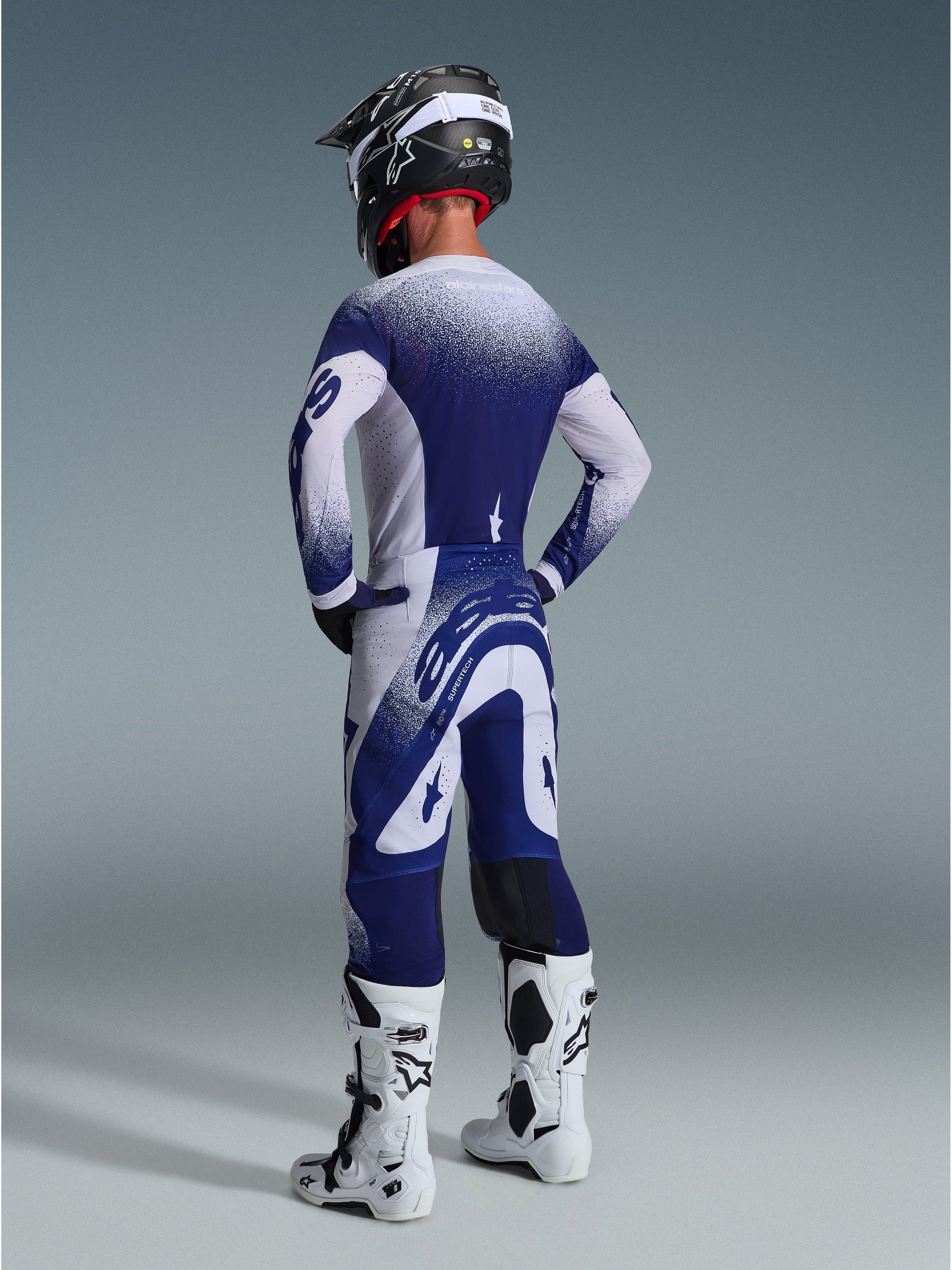 Alpinestars 2026 Supertech Scenz broek, motocrossbroek, lichtgrijs en donkerblauw, achteraanzicht gedragen met bijpassende jersey en witte laarzen, voorzien van stippengrafieken met kleurverloop en een sportieve atletische pasvorm voor off-road racing