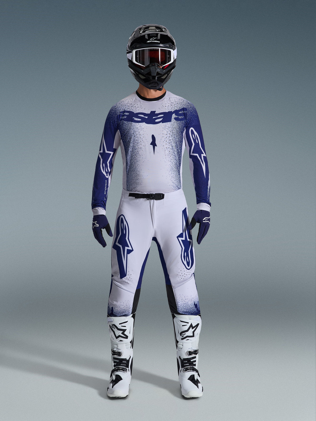 Motorcrosser in de Alpinestars 2026 Supertech Scenz Gear Set, wit en donkerblauw jersey met bijpassende broek, voorzien van grote Astars-logo's en een gespikkeld grafisch ontwerp, gecombineerd met een zwarte helm, wit-zwarte beschermende laarzen en bijpassende blauwe handschoenen, complete professionele race-outfit