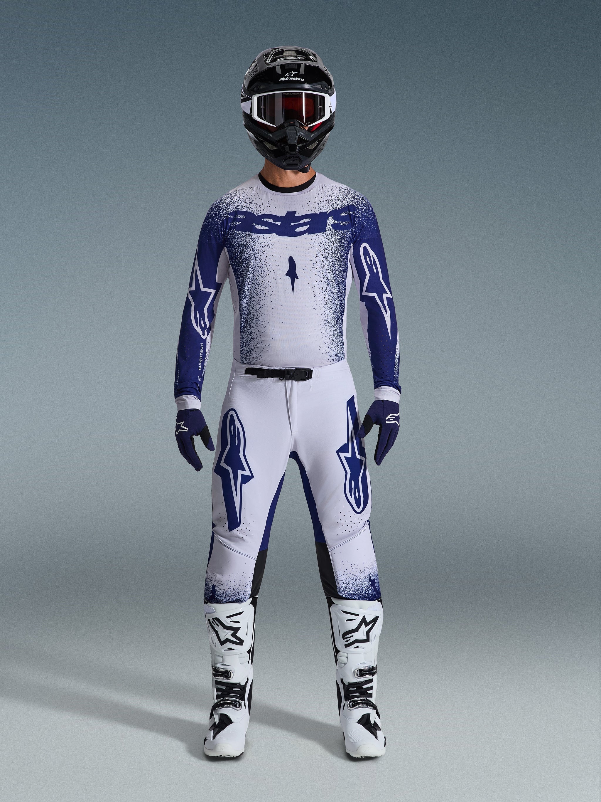 Motorcrossrijder in Alpinestars 2026 Supertech Scenz-kledingset, lichtgrijs en donkerblauw, inclusief jersey en broek met bijpassende blauwe handschoenen, witte en zwarte laarzen, en een zwarte helm met witte crossbril, gecoördineerde outfit voor professionele off-road racing