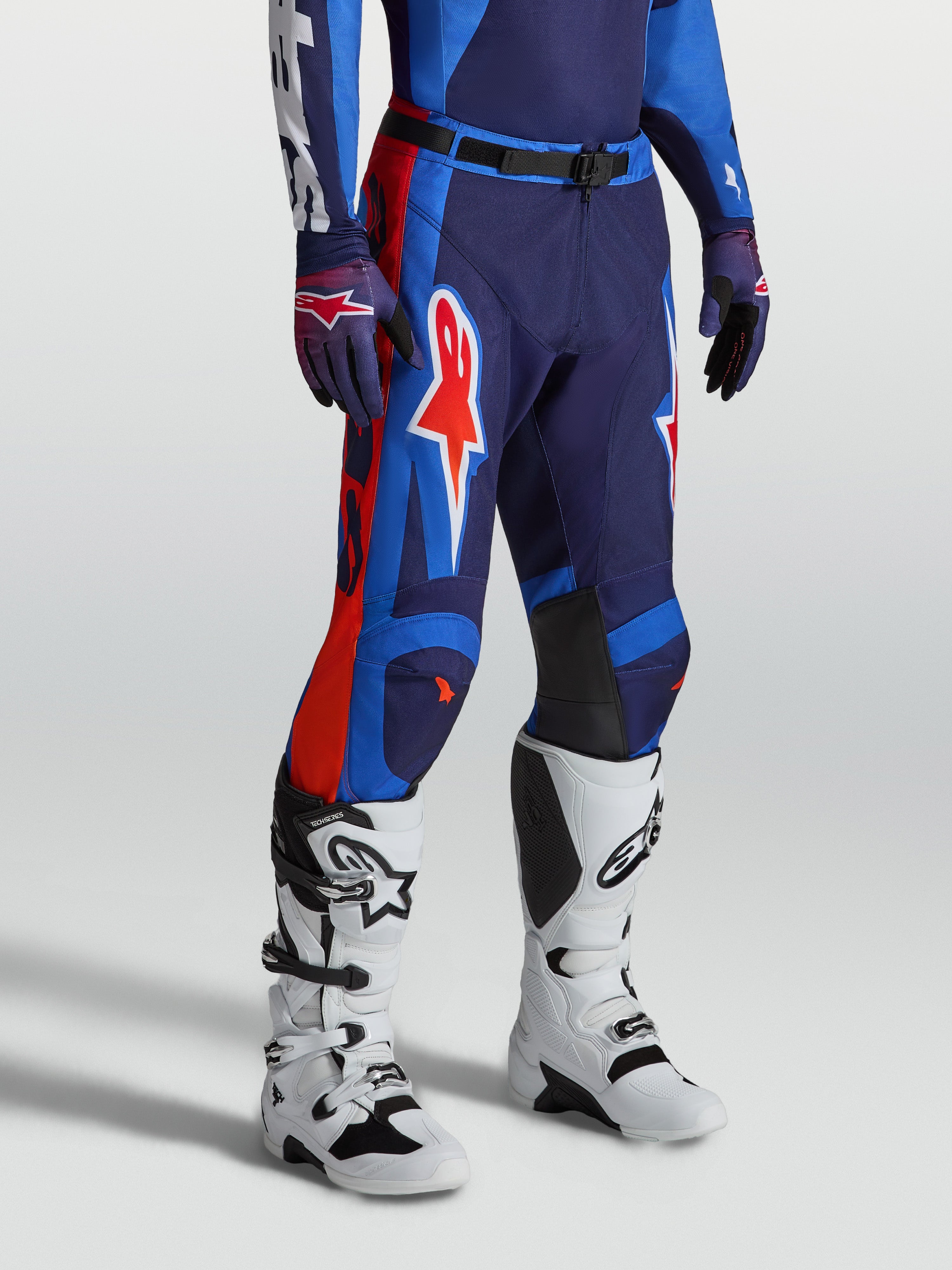 Alpinestars Racer Hunt broek, heren motorcrossbroek, blauw en vuurrood, duurzaam 600D polyweefsel met ergonomische voorgevormde racepasvorm, gedragen met witte motorlaarzen.