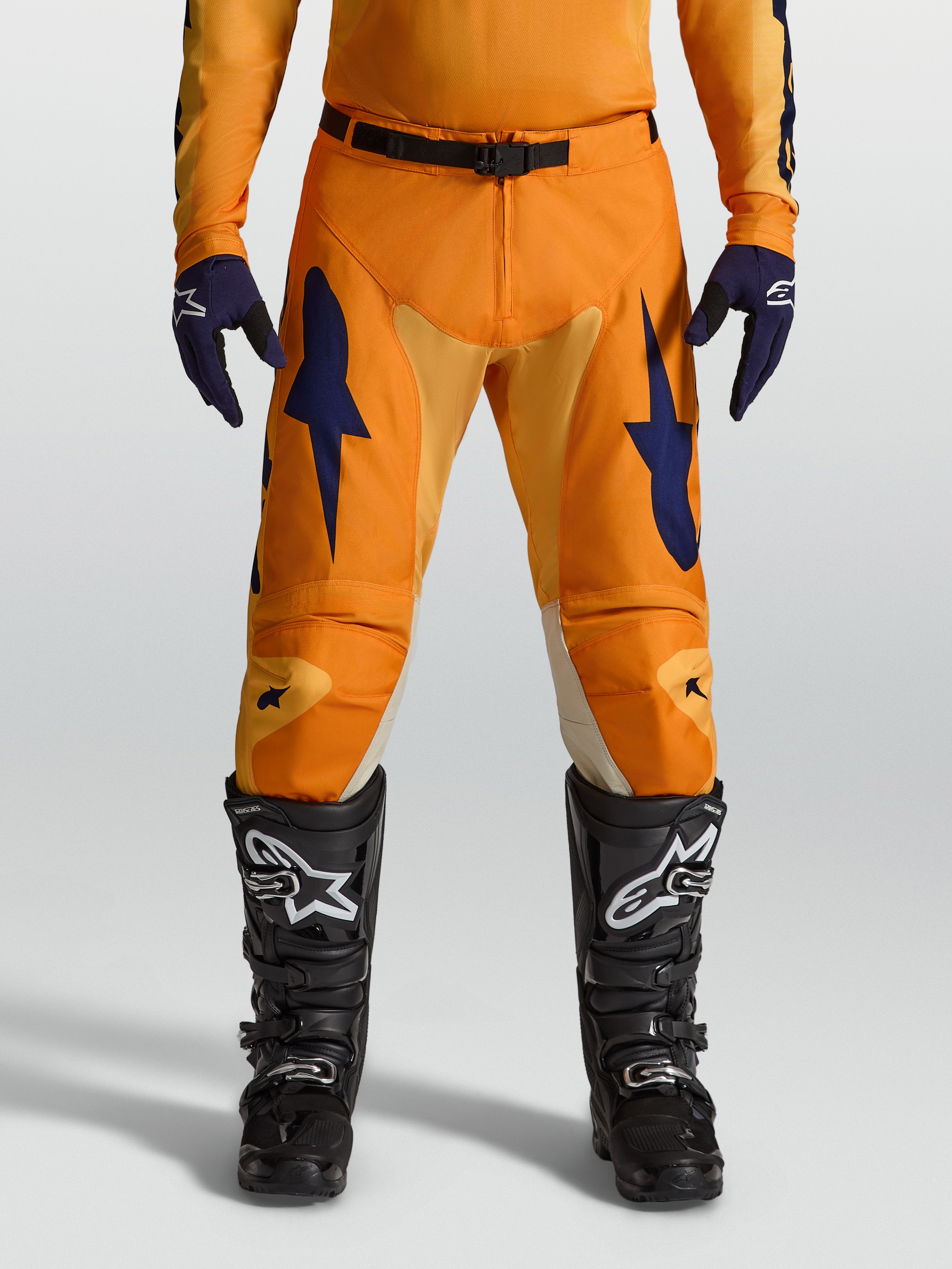 Alpinestars Racer Amber Broek, motocrossbroek voor heren, oranje en donkerblauw, 600D polyfabric met stretchinzetstukken, racepasvorm voor off-road rijden, gedragen met zwarte motorlaarzen.