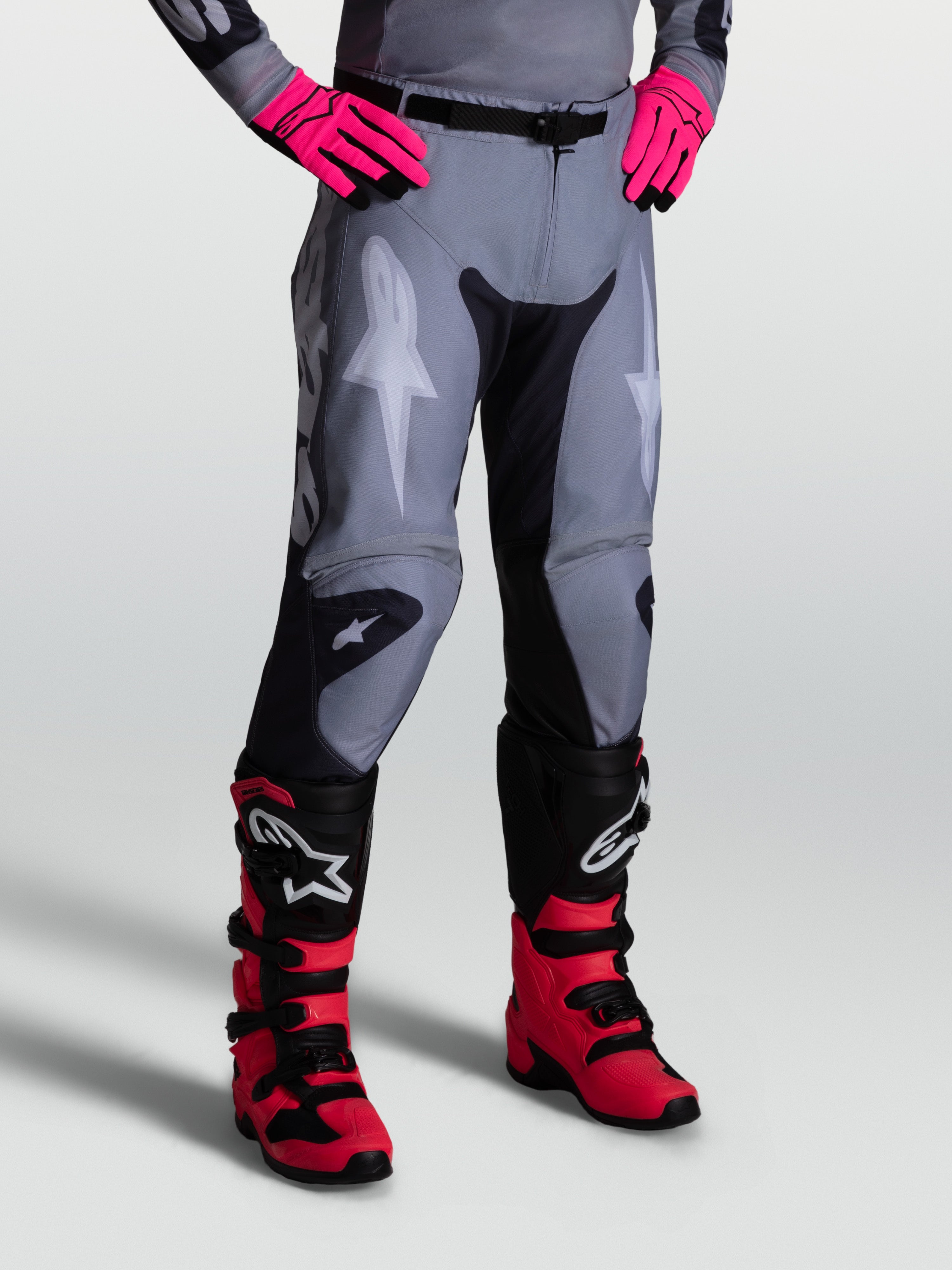Alpinestars Racer Amber broek, herenmotocrossbroek, grijs en zwart, duurzaam 600D polyweefsel met stretchpanelen, gedragen met roze handschoenen en zwarte crosslaarzen.