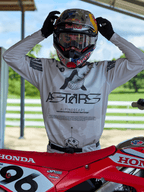 Motocrossrijder in Alpinestars 2025 Racer Hollow-uitrusting, lichte camo jersey met opvallende ASTARS-branding, stelt een Red Bull-helm met heldere crossbril af, staand naast een rode Honda crossmotor in een off-road omgeving.