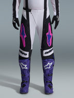 Motocross rider wearing the Alpinestars 2026 Techstar Knif Gear Set, white and black pants with pink and blue logos, purple boots with white branding, black gloves, featuring an athletic race fit and laser-cut ventilation for off-road racing
