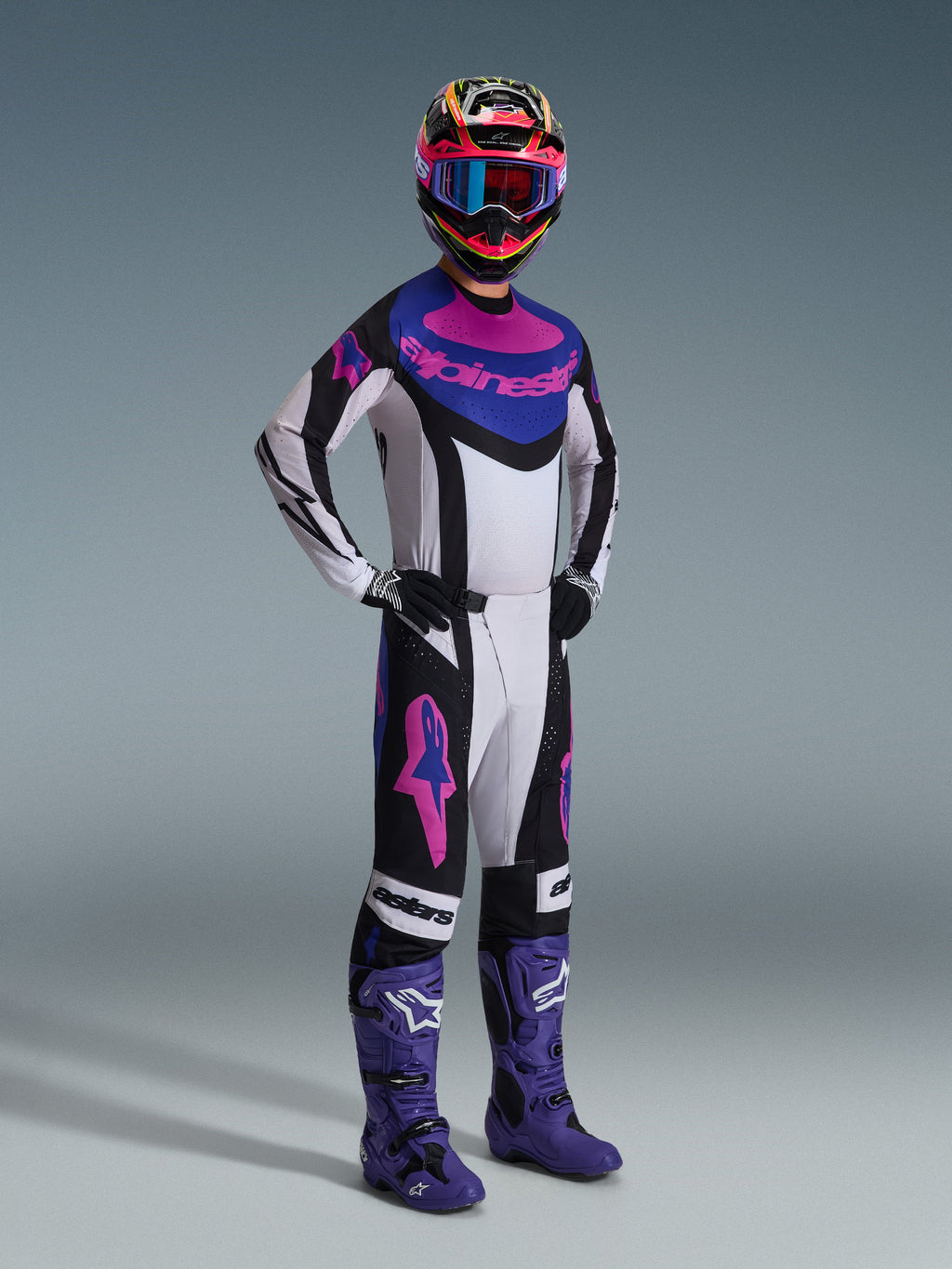 Motocross rider wearing the Alpinestars 2026 Techstar Knif Gear Set, white jersey with black, blue, and pink accents, matching pants with pink Astars logos, multi-colored helmet with goggles, purple racing boots, coordinated high-performance gear for motocross competition