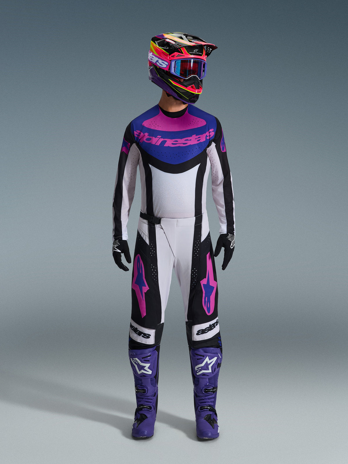 Alpinestars 2026 Techstar Knif Gear Set, complete motorcross-uitrusting, wit, zwart, paars en roze, gedragen met een veelkleurige helm, blauwe motorcrossbril, paarse laarzen en zwarte handschoenen, gecoördineerde hoogwaardige set voor off-road racing