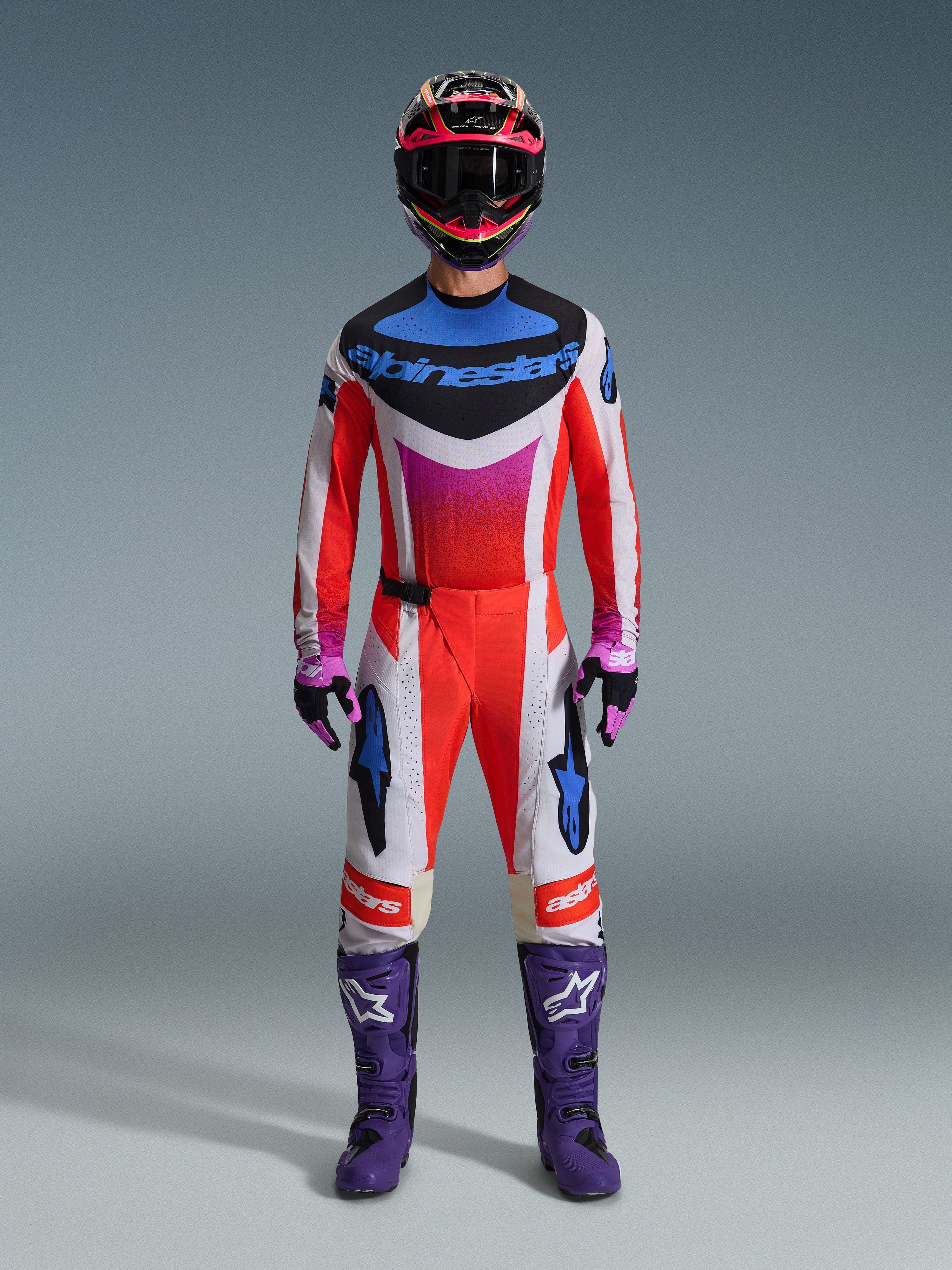 Motorcrosser die de 2026 Techstar Knif Gear Set draagt, compleet racetenue in rood, wit en zwart met blauwe merkdetails en roze accenten, met paarse laarzen met witte logo's, roze en zwarte handschoenen en een zwart-roze helm, professionele off-road race-uitrusting