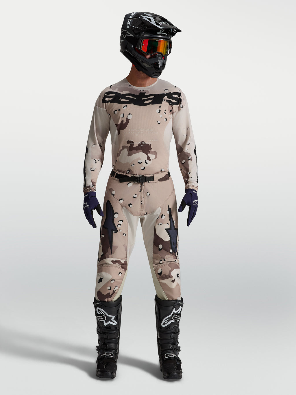 Alpinestars Racer Lone broek en jersey, heren motorcross-set in desert camo zwart, gedragen door model met zwarte motorlaarzen en helm, professionele off-road race-uitrusting.