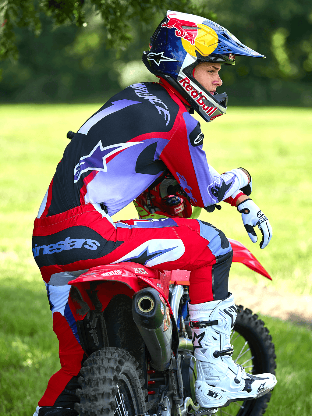 Alpinestars 2025 Fluid Wurx crossbroek in paars en felrood, motorcrosser zittend op een rode crossmotor, bijpassende jersey, wit-zwarte Tech 10 laarzen, witte handschoenen en Red Bull-helm, complete off-road race-outfit in een buitenomgeving.