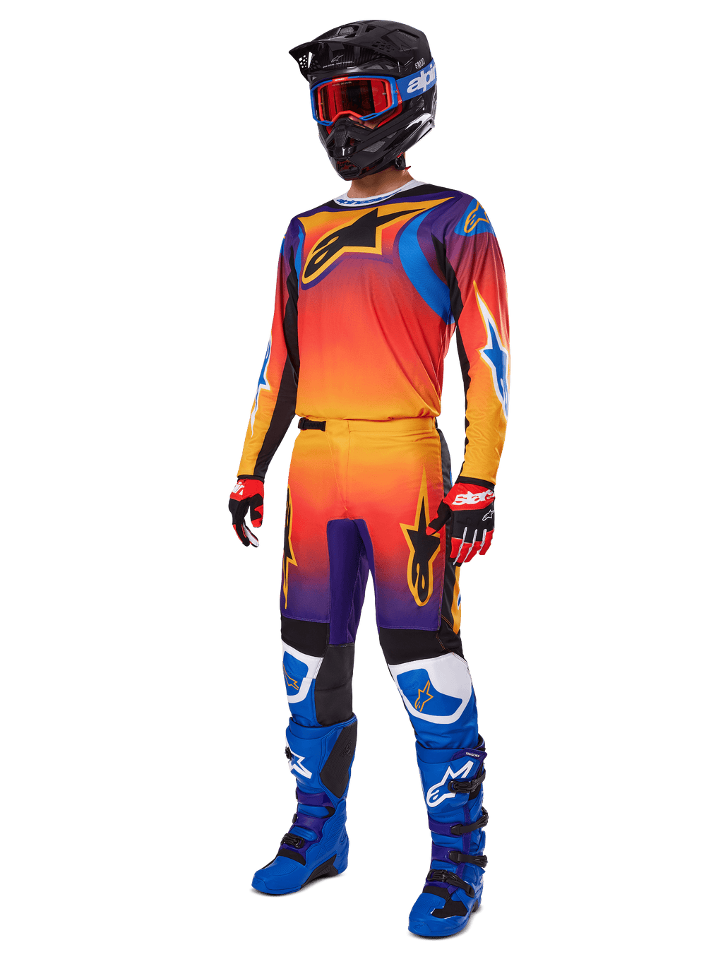 Motorcrosser in volledige race-uitrusting, Alpinestars 2025 Fluid Wurx broek en bijpassende jersey met een multicolor verloop van oranje, rood en paars, zwarte helm met rode crossbril, blauwe en zwarte laarzen, rode en zwarte handschoenen, gecoördineerde outfit voor off-road racing
