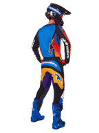 Motorcrosser van achteren gezien, Alpinestars 2025 Fluid Wurx broek, multicolor ontwerp met blauwe, oranje, gele en paarse accenten, gecombineerd met een bijpassende jersey, felblauwe laarzen en zwarte helm, complete off-road race-uitrusting