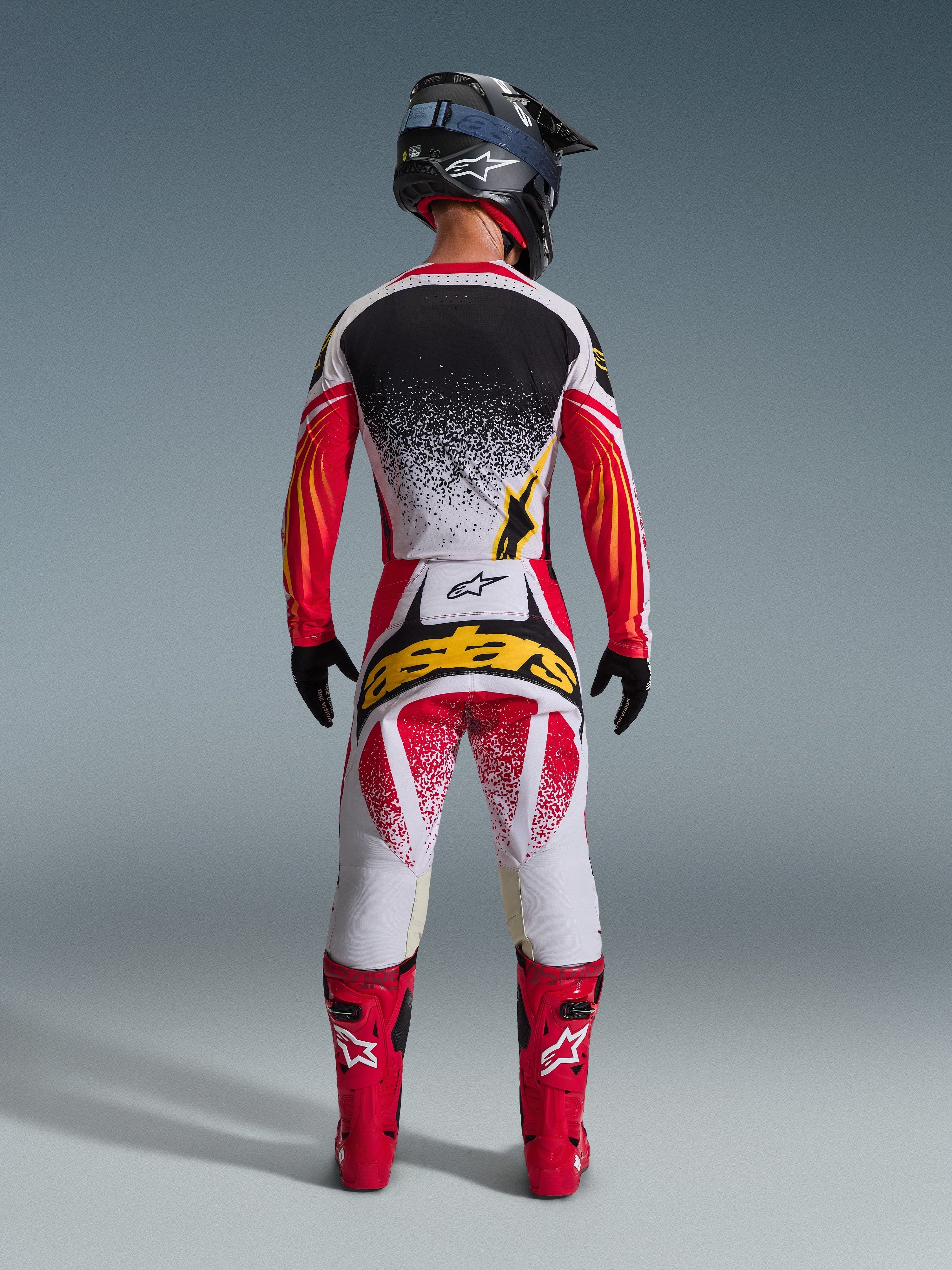 Rear view of a motocross rider wearing the Alpinestars 2026 Techstar Nomur Jersey in Black, Red, and White with yellow accents, paired with matching motocross pants featuring bold yellow branding, bright red racing boots, and a black helmet, complete coordinated gear set for off-road racing.