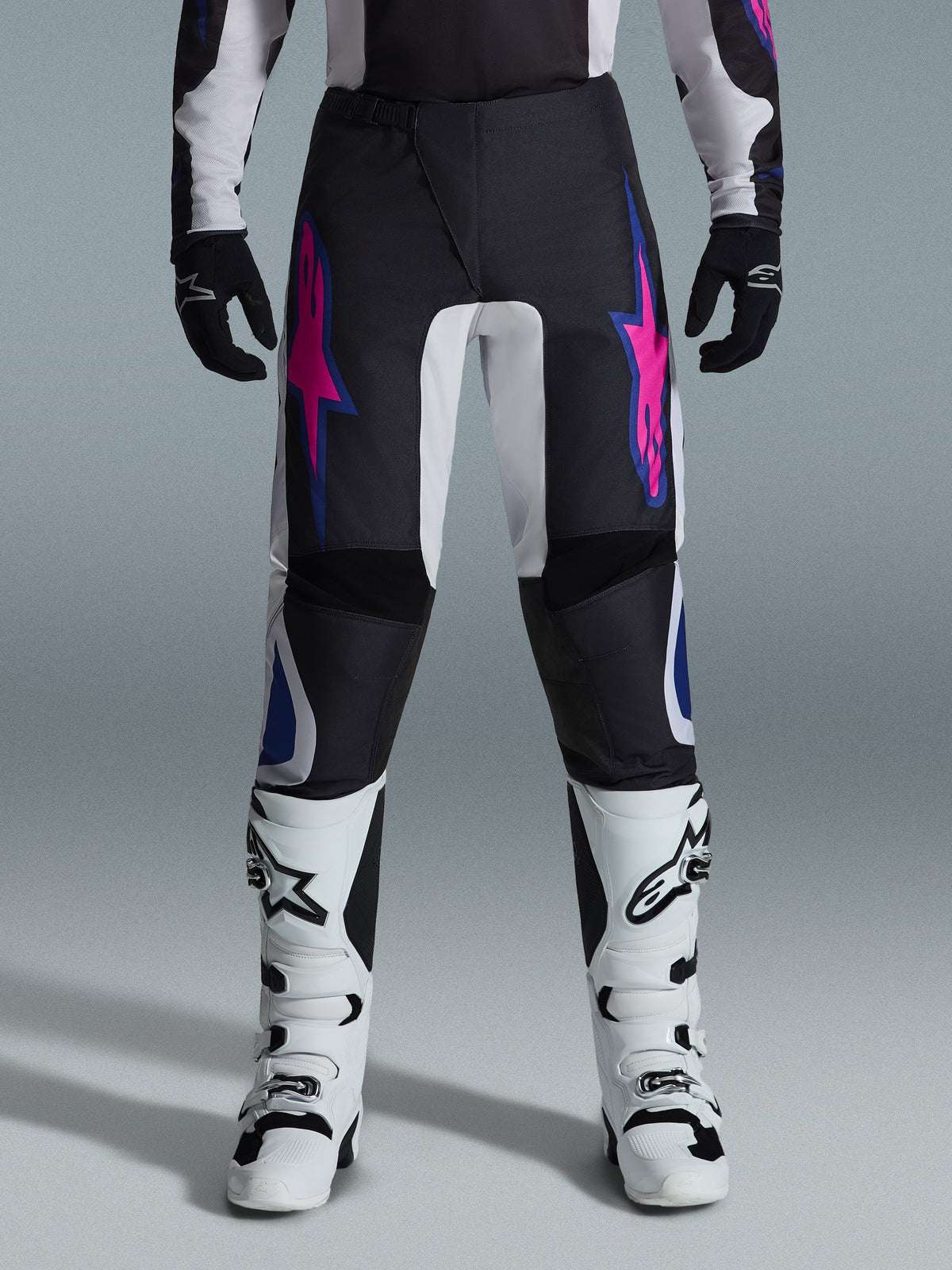 Alpinestars 2026 Fluid Grid Pants, Motocross Pants, Black with Purple and Blue Star Designs, worn with white motocross boots and black gloves, featuring reinforced panels and a sporty off-road fit, gray background