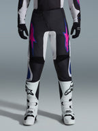 Alpinestars 2026 Fluid Grid Pants, Motocross Pants, Black with Purple and Blue Star Designs, worn with white motocross boots and black gloves, featuring reinforced panels and a sporty off-road fit, gray background