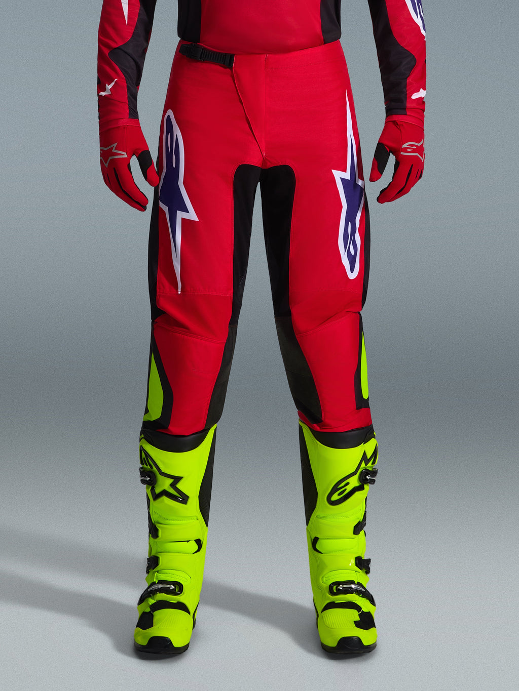 Alpinestars 2026 Fluid Grid broek, motorcross broek, rood met zwarte panelen en neongele accenten, voorzien van grote witte en paarse ster-logo's op de dijen, gedragen met rode handschoenen en neongele laarzen, duurzame constructie voor off-road racing