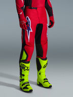 Alpinestars 2026 Fluid Grid crossbroek, motorcrossbroek, rood, zwart en paars, gedragen met neongele laarzen en rode handschoenen, met zwarte panelen en een groot paars-wit sterlogo, duurzaam sport-fit ontwerp voor off-road racen