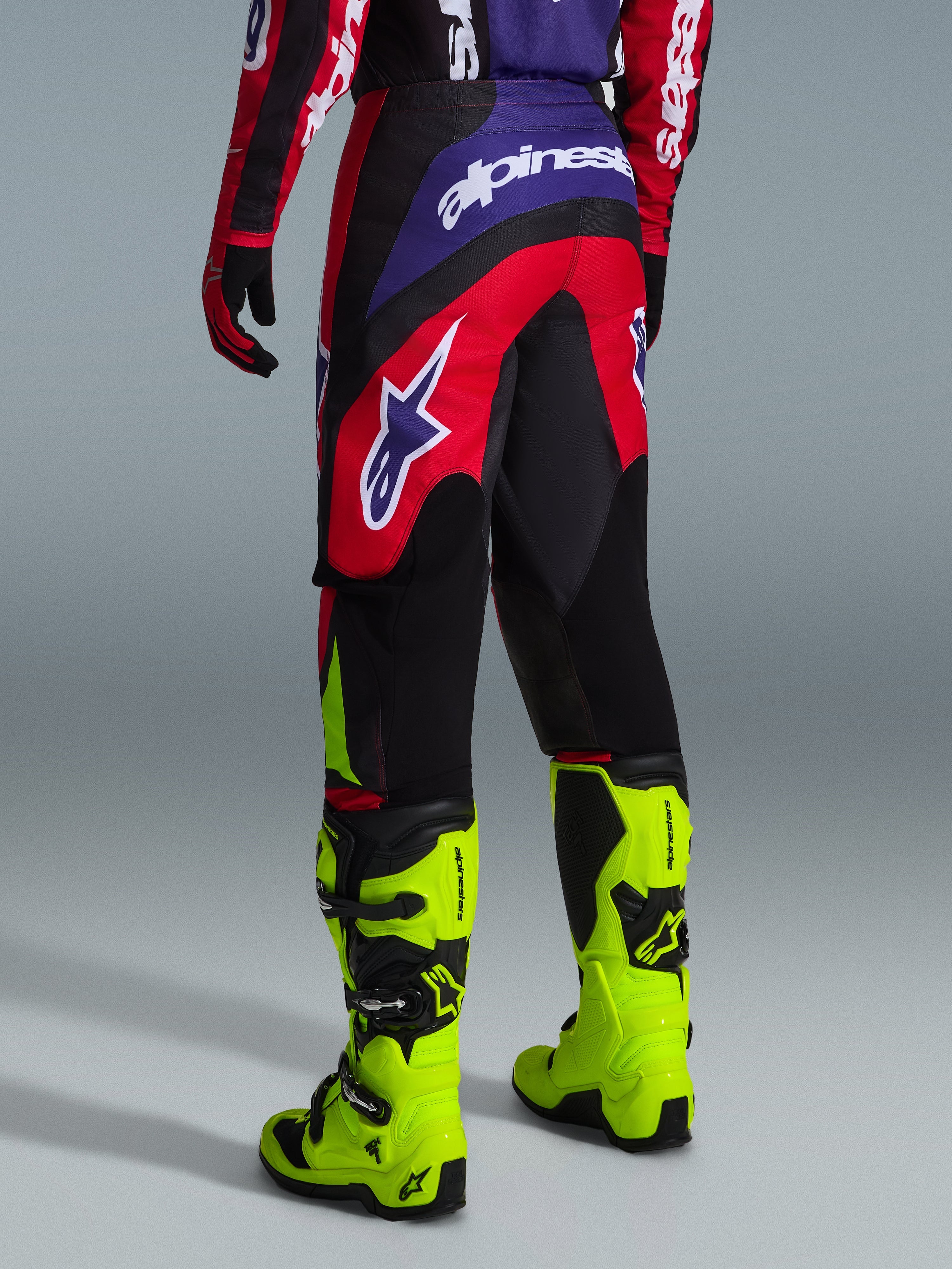 Alpinestars 2026 Fluid Grid crossbroek, motorcross broek, rood, zwart en paars, achteraanzicht met groot wit logo, gedragen met neongele racelaarzen en bijpassende jersey, duurzaam sport-fit ontwerp voor off-road prestaties