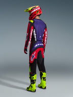 Alpinestars 2026 Fluid Grid Pants, motorcrossbroek, rood, zwart en paars, van achteren gedragen door een rijder met bijpassende jersey en fluogele laarzen, met opvallende branding en ergonomisch ontwerp voor off-road prestaties