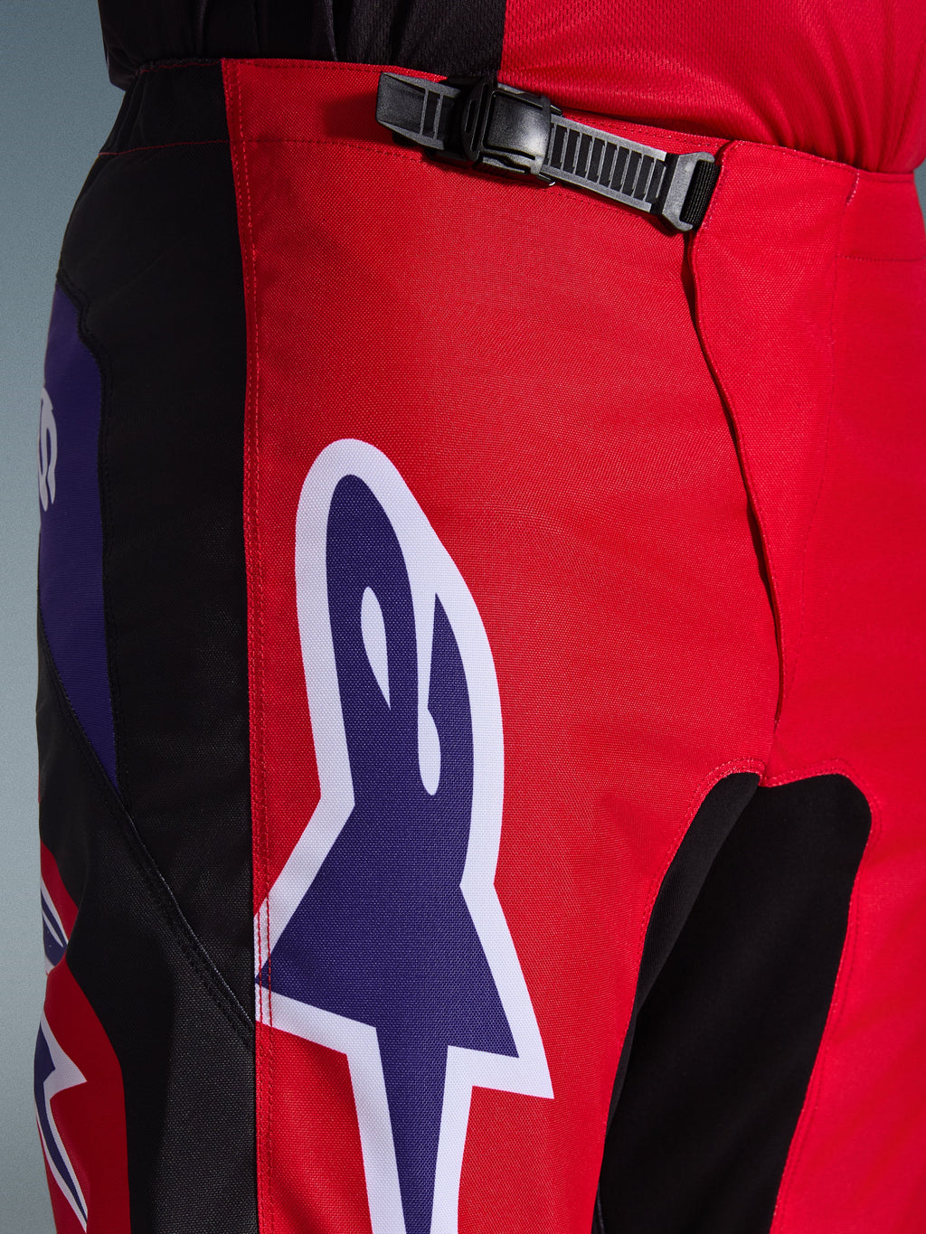 Alpinestars 2026 Fluid Grid broek, motorcross broek, rood, zwart en paars, close-up van de taille en heupen met een zwart ratelsluitingssysteem en een groot paars Alpinestars sterlogo met witte omlijning, duurzame getextureerde stof voor off-road racen.