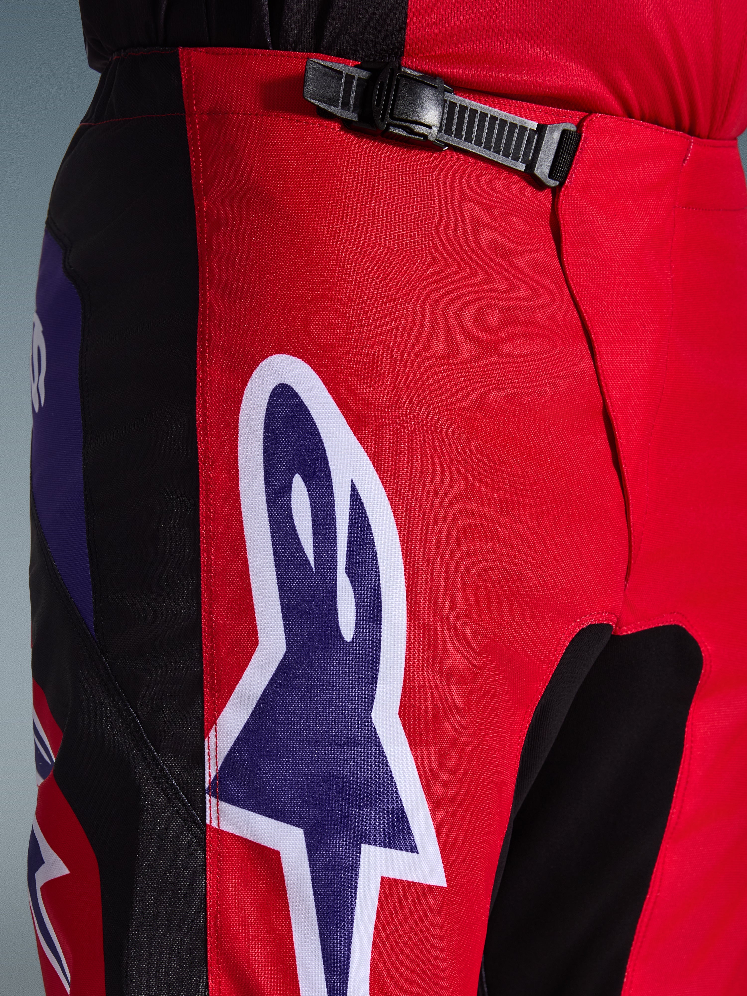 Alpinestars 2026 Fluid Grid broek, motorcross broek, rood, zwart en paars, close-up van de taille en heupen met een zwart ratelsluitingssysteem en een groot paars Alpinestars sterlogo met witte omlijning, duurzame getextureerde stof voor off-road racen.