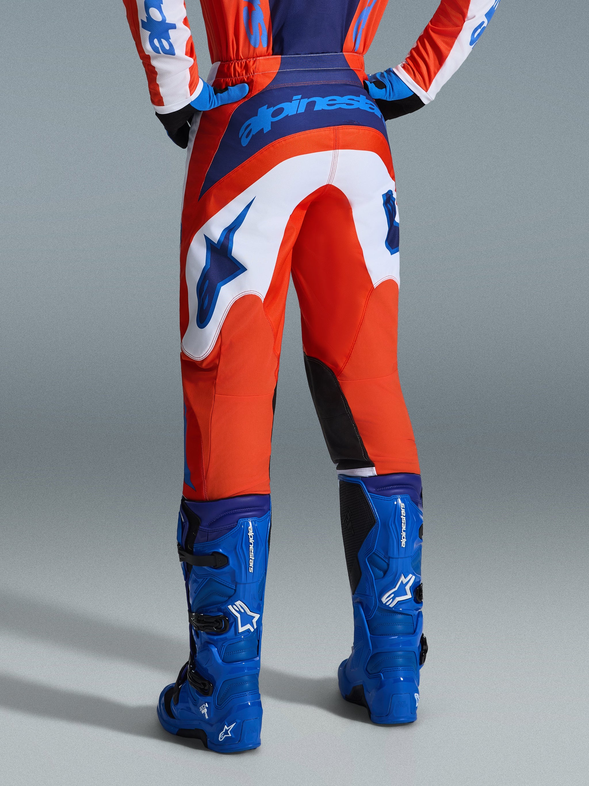Alpinestars 2026 Fluid Grid Pants, Motocross Pants, Orange and UCLA Blue with white panels, rear view showing bold blue branding on the seat and star logo, paired with matching blue motocross boots, professional off-road racing gear