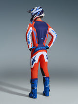 Motocross rider from behind wearing Alpinestars 2026 Fluid Grid Pants in Orange and UCLA Blue, paired with a matching jersey, blue racing boots, and a blue and white helmet, full gear set for off-road riding.