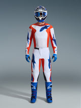 Motocross rider wearing complete racing gear, Alpinestars 2026 Fluid Grid Pants in Orange and UCLA Blue, white jersey with orange sleeves and blue logos, matching blue helmet and goggles, blue boots and blue gloves, coordinated outfit for off-road racing