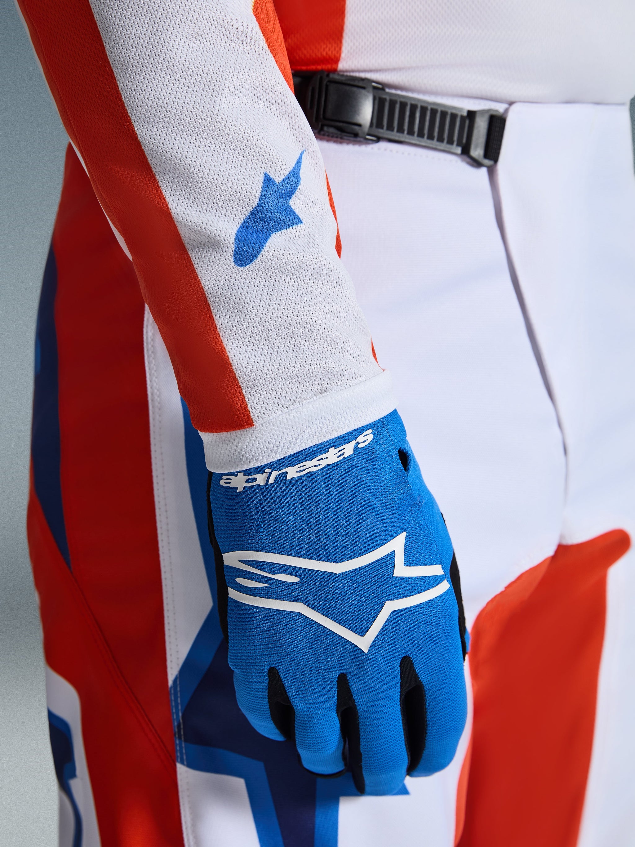 Alpinestars 2026 Fluid Grid Pants, Motocross Pants, Orange and UCLA Blue, close-up featuring a matching blue glove and white mesh jersey sleeve with blue logos, showcasing the ratchet waist closure and athletic fit.