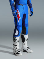 Alpinestars 2026 Fluid Grid Pants, Motocross Pants, Blue and Red, worn with white motocross boots and blue gloves, featuring a large red star logo on the thigh and black inner knee panels, designed for off-road riding.