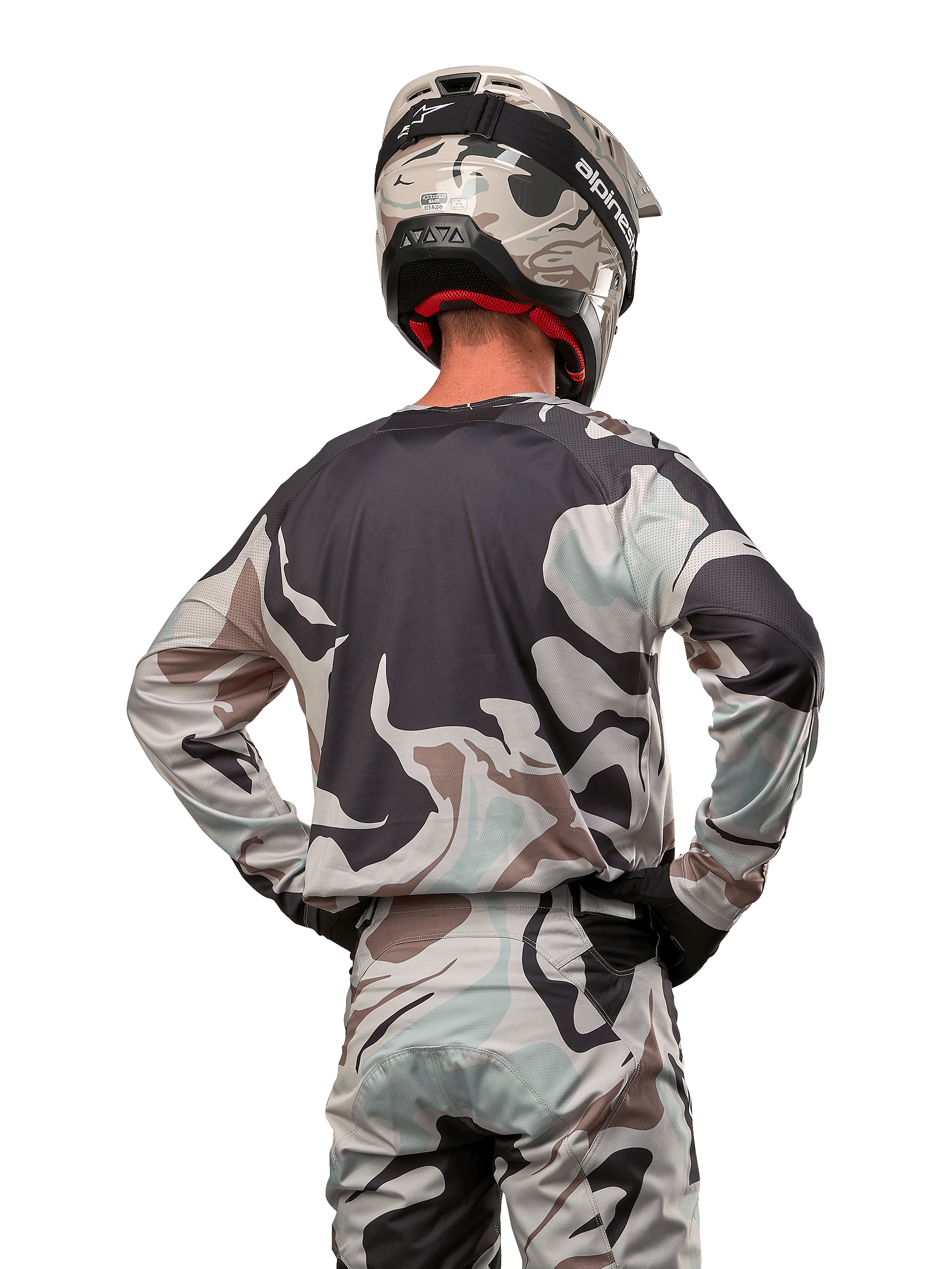 Motocrossrijder van achteren gezien in de Alpinestars 2024 Racer Tactical Jersey in Iron Camo Dust Gray, bijpassende camouflagebroek en helm, handen op de heupen, gecoördineerde off-road race-uitrusting