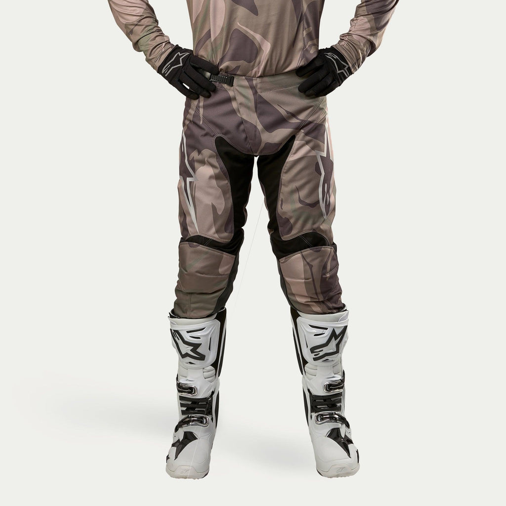 Alpinestars 2024 Racer Tactical broek, motorcrossbroek, Military Green Camo Brown, met grote witte Alpinestars-logo's op de dijbenen en zwarte panelen aan de binnenkant van de benen, gedragen met wit-zwarte motorcrosslaarzen en zwarte handschoenen, bijpassende camouflage jersey, vooraanzicht.