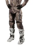 Alpinestars 2024 Racer Tactical broek, motorcross broek, militair groen camouflagebruin, gedragen met bijpassende camouflage jersey, zwarte handschoenen en witte laarzen met ster-logo's, voorzien van zwarte stretchpanelen en een ergonomisch ontwerp, off-road outfit op een witte achtergrond
